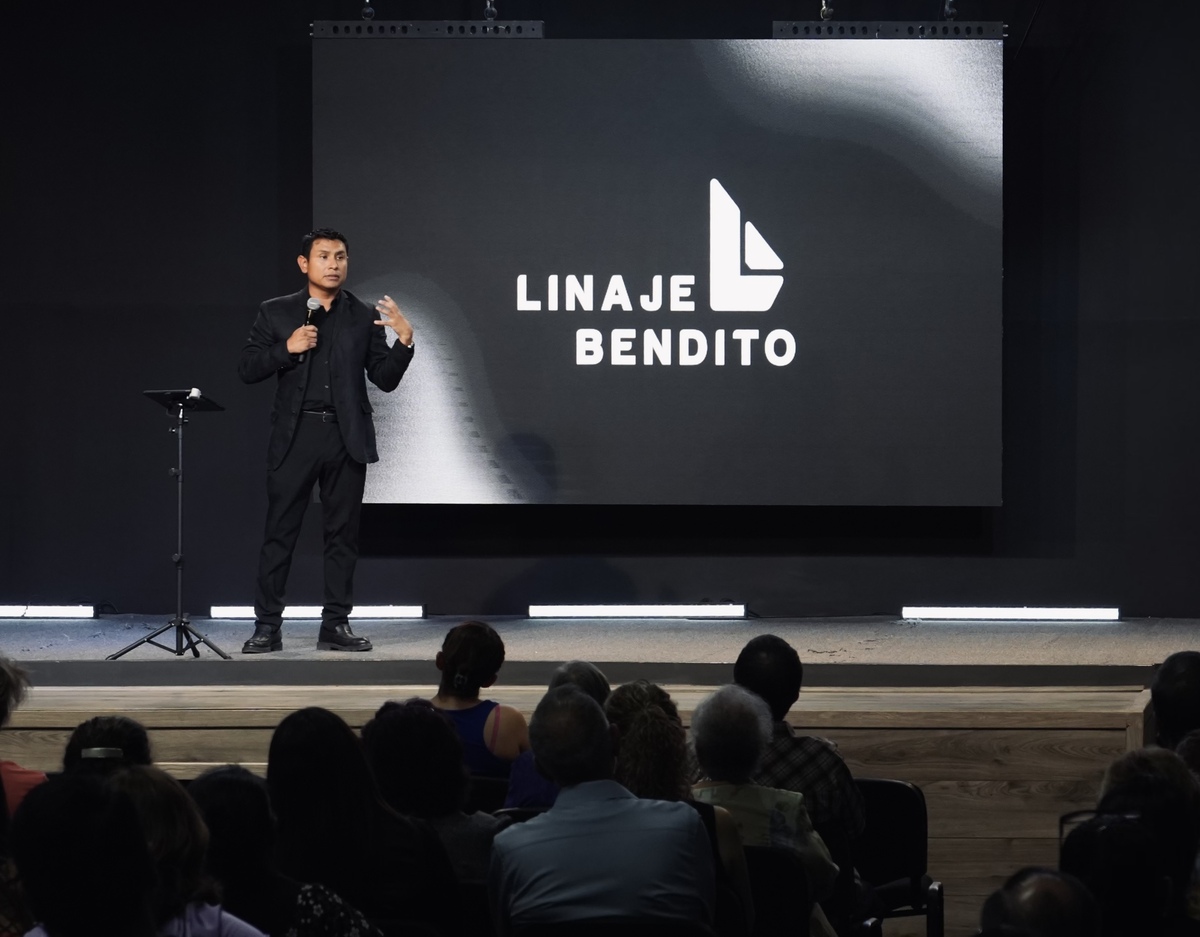 El Pastor Iván predicando a la congregación desde el escenario, con el logotipo de la iglesia de fondo